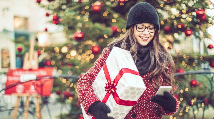 woman in hat and coat carrying a white package with a red ribbon checking her cell phone